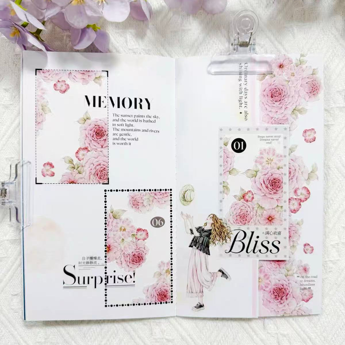 An open handmade scrapbook journal or planner binder with floral pink and white pages, decorative labels, and text including 'MEMORY', 'Surprise!', and 'Bliss', styled with purple flowers at the top on a white textured fabric background.