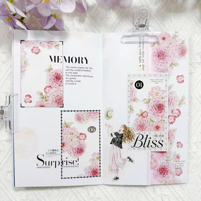 An open handmade scrapbook journal or planner binder with floral pink and white pages, decorative labels, and text including 'MEMORY', 'Surprise!', and 'Bliss', styled with purple flowers at the top on a white textured fabric background.